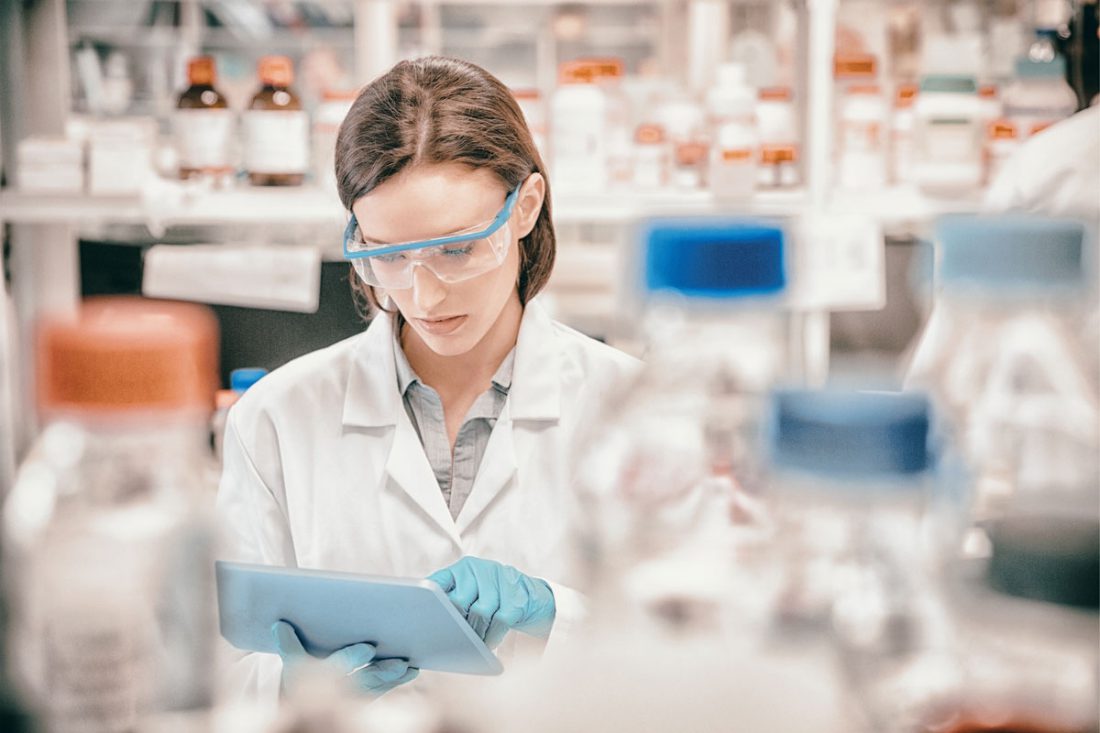 A chemist in a lab surrounded by bottles. They are wearing safety glasses and blue gloves, and they are using a tablet.